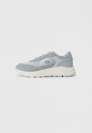 Zapatilla Lacoste en gris claro y blanco con cordones en la parte delantera, suela acolchada y logo de cocodrilo bordado en el lateral, mostrada de perfil.