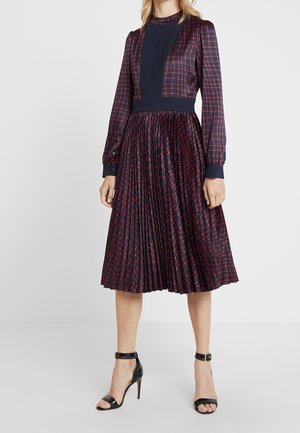 Vrouw draagt een marineblauwe jurk met rood geruit patroon, lange mouwen, geplooide rok en zwarte sandalen met hoge hak en enkelriem.