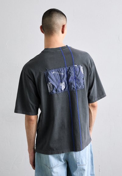 Gray oversized t-shirt with short sleeves, featuring a blue stitched patch on the back, adorned with white graphic designs.