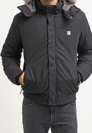 Person trägt eine schwarze Winterjacke mit Reißverschluss und einem mit Fell gefütterten Kapuzen, die Hände in den Vorderentaschen, kombiniert mit dunklen Jeans.