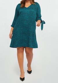 Teal klänning med svart prickmönster, v-ringning, trekvartslånga ärmar och knytdetaljer på ärmarna. Bärs med svarta spetsiga klackskor.