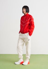 Rød vindjakke med hvite accenter, kremfargede joggebukser og hvite sneakers med røde detaljer. Glatt tekstur og atletisk design.