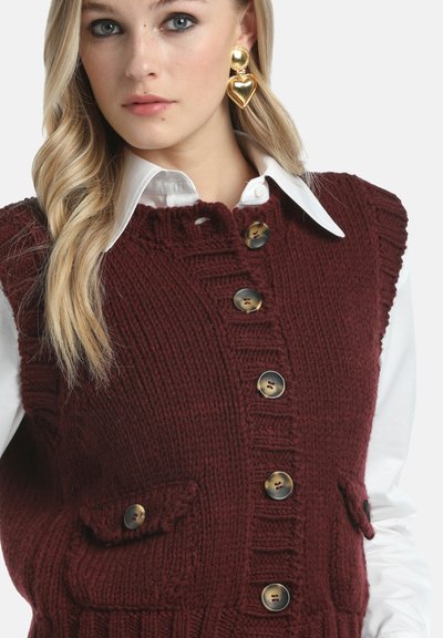 Blond kvinna som bär stora guldhjärtformade örhängen, vit skjorta med krage och vinröd stickad väst med knappar och fickor.