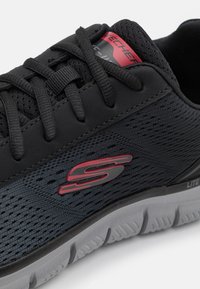 Black athletic shoe featuring a mesh fabric upper, 3D-patterned texture, red logo accent, and a cushioned white sole with textured grooves.