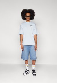 Lyseblå oversized t-shirt med kortærmede ærmer og logo, parret med knælange denim shorts. Sorte og hvide sneakers supplerer stribede sokker.