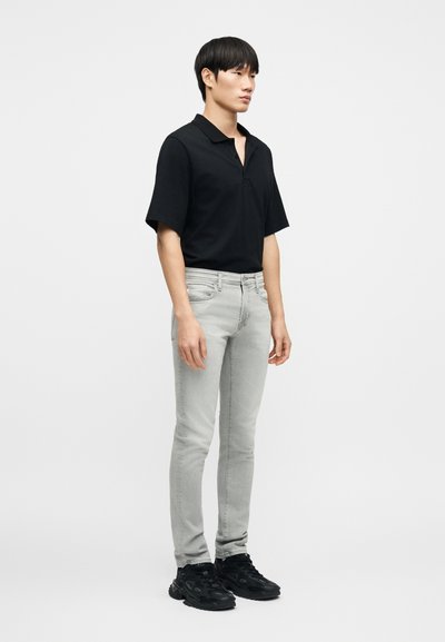 Μαύρο πόλο μπλουζάκι με κοντά μανίκια και άνετη εφαρμογή. Ανοιχτό γκρι τζιν παντελόνι στενής γραμμής με διακριτική ξεθώριασμα. Μαύρα αθλητικά παπούτσια με υφασμένη σόλα.