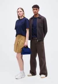 Γυναίκα με μπλε μπλούζα, φούστα με κίτρινο καρό σχέδιο, λευκά αθλητικά παπούτσια, κρατώντας μπλε τσάντα· άνδρας με σκούρο σακάκι, μπλε μπλούζα, καφέ παντελόνι, στέκονται δίπλα δίπλα.