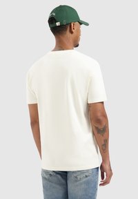 Crème t-shirt met korte mouwen, gemaakt van zachte stof, met een klassieke snit. G gedragen met lichtblauwe spijkerbroek en een groene pet met geborduurde details.