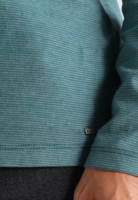 Türkise Langarmbluse mit einem strukturierten, gestreiften Muster. Verfügt über ein kleines metallisches Etikett am Saum. Der Stoff wirkt weich und leicht dehnbar.