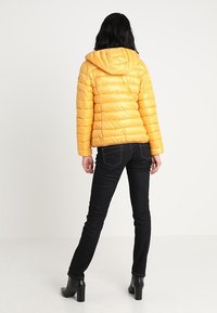 Femme aux cheveux noirs portant une doudoune jaune à capuche, un jean skinny noir et des bottines à talons noires, se tenant debout, de dos face à l'avant.