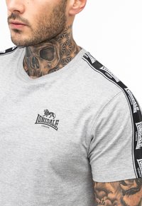 Tricou scurt gri cu mânecă scurtă, având logo-ul Lonsdale pe piept. Benzi negre cu text alb pun în evidență umerii. Textură netedă din bumbac.