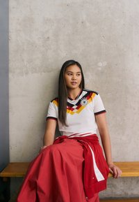 Jonge vrouw in een Duits voetbalshirt en rode rok die op een houten bank tegen een betonnen muur zit, en opzij kijkt.