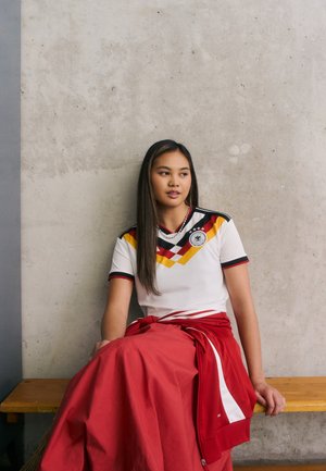 Tânără femeie îmbrăcată într-un tricou de fotbal al Germaniei și o fustă roșie, stând pe o bancă de lemn lângă un perete de beton, uitându-se în lateral.