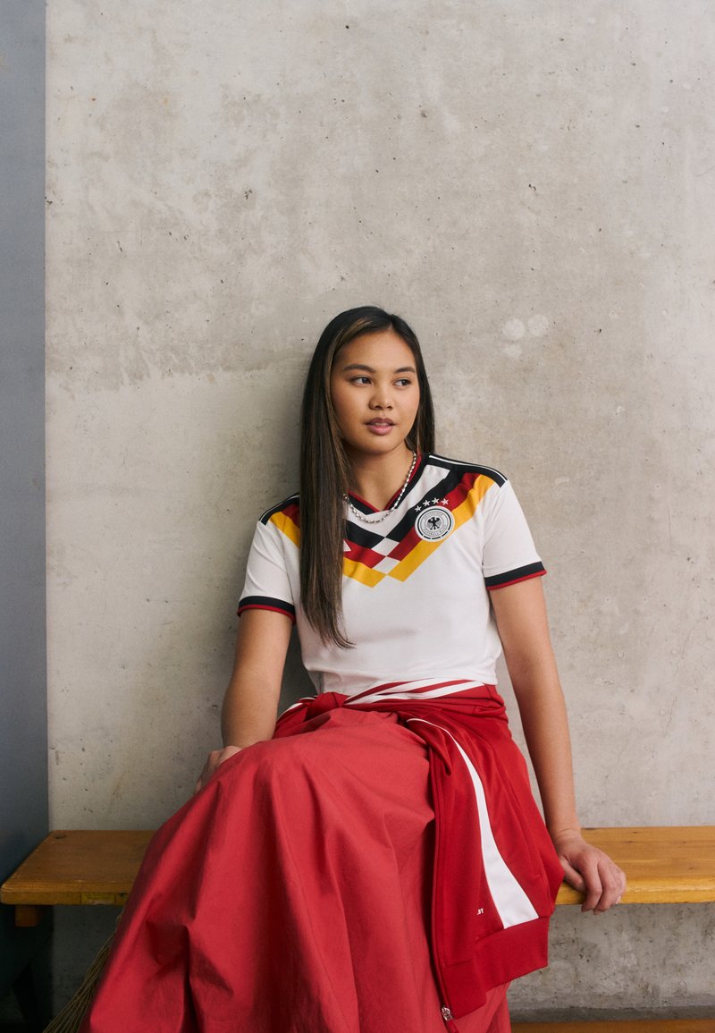 Jonge vrouw in een Duits voetbalshirt en rode rok die op een houten bank tegen een betonnen muur zit, en opzij kijkt.
