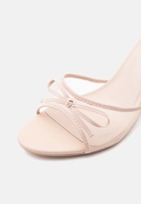 Μπεζ σανδάλι με ανοιχτά δάχτυλα και ψηλό τακούνι, με διάφανα πλεκτά λουριά και διακοσμητικό φιόγκο στο μπροστινό λουρί.