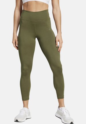 Frau trägt olivgrüne Leggings mit hoher Taille und weiße Sportschuhe, steht mit leicht gespreizten Beinen vor einfachem Hintergrund.