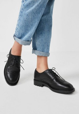 Černé kožené brogue boty s perforovanými detaily, šněrování a chunky texturovanou podrážkou, spárovány s vyhrnutými světle modrými džíny.