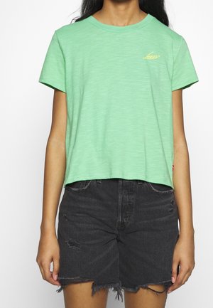 Zielony krótki t-shirt z miękkiego materiału z krótkimi rękawami, ozdobiony małym żółtym logo na lewej piersi. Noszony z czarnymi szortami jeansowymi.