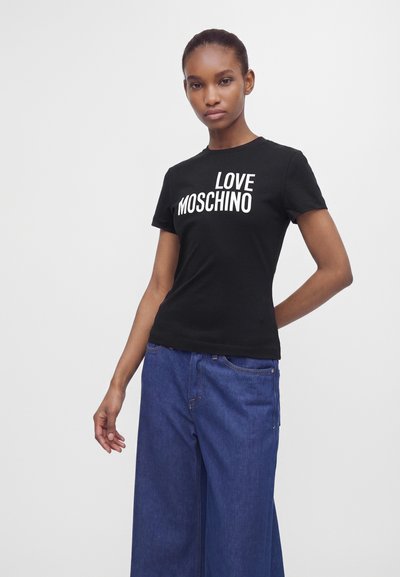 Млада жена, носеща черна тениска "LOVE MOSCHINO" и широки сини дънки, стояща с една ръка зад гърба си пред едноцветен фон.