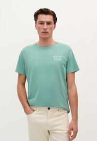 Mintgroene crew neck t-shirt van zachte stof, met de opdruk "Lifeguard's Club" op de linkerkant van de borst, gecombineerd met lichtgekleurde broek.