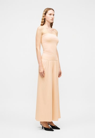 Γυναίκα που στέκεται, φορώντας ένα μπεζ τοπ χωρίς τιράντες και παντελόνι φαρδιάς γραμμής στο ίδιο χρώμα, σε συνδυασμό με μαύρες γόβες με μυτερή μύτη, μπροστά σε ένα απλό φωτεινό φόντο.