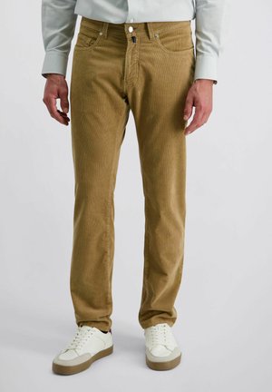 Man in een lichtbruine corduroy broek, witte sneakers en een lichtgrijze lange mouwen shirt, staande tegen een effen achtergrond.