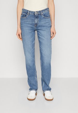 Person iført lyseblå jeans med lige ben, hvide sneakers med brune såler og en hvid top stoppet ind, stående mod en ensfarvet baggrund.