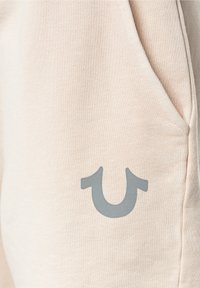 Jasnobeżowe dresy o gładkiej fakturze, wyposażone w przednią kieszeń i szare logo po boku.