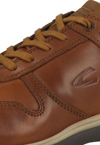Brown leather sneaker with beige laces, curved stitching details, subtle logo embossed on the side, and a cream-colored outsole.