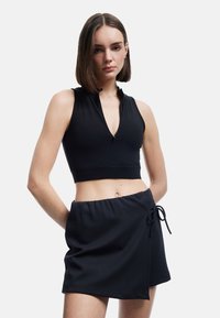 Mujer con cabello castaño liso que lleva un top corto negro sin mangas con cremallera frontal y una mini falda negra envolvente con cordón.