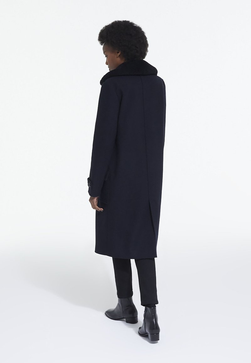 Manteau en laine bleu marine avec un col haut, des poches latérales et une fente au dos, associé à un pantalon noir et des bottes chelsea. Texture lisse et design structuré.