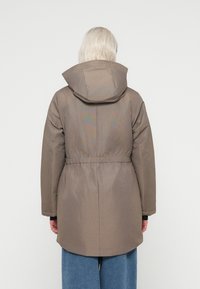 Podloženo zunanje oblačilo v taupe odtenku, ki ima kapuco, elastični pas in zapestne manšete ter daljšo zasnovo na hrbtu. Gladka tekstura blaga.