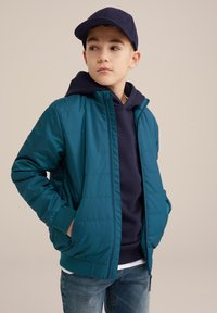 Teal puffig jacka med dragkedja, ribbade ärmslut, bärs över en marinblå hoodie. Blå keps, ljusa jeans. Slät struktur, kviltat mönster.