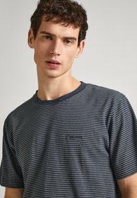 Camiseta de algodón a rayas en azul marino y blanco, con cuello redondo y mangas cortas; presenta un pequeño logo en el lado izquierdo del pecho.