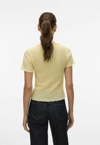 Kortermet gul topp laget av strukturert strikket stoff, med en bølget nederkant og en tettsittende fasong, brukt med mørke jeans.
