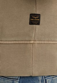 Beige gebreide sweatshirt met ribbeltextuur, voorzien van een zwart label met de tekst "PME LEGEND," geplaatst bovenop blauwe denim stof.