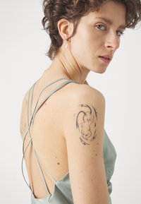 Mintgrön satin klänning med låg rygg och korsade band. Har en synlig tatuering på axeln. Strukturerad tyg, slät yta.