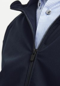 Veste zippée marine en tissu lisse, avec une fermeture éclair noire et une chemise à col bleue et blanche rayée en dessous.