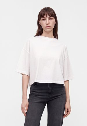 Vrouw met stijl bruin haar, die een losse witte korte mouwen blouse en een hooggetailleerde donkergrijze spijkerbroek draagt, staand tegen een effen achtergrond.