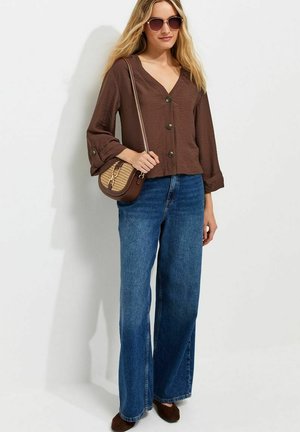 Vrouw draagt een bruine blouse met knopen, wijde blauwe jeans, bruine zonnebril en draagt een kleine bruine gevlochten schoudertas.