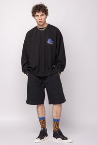 Sudadera negra de gran tamaño con gráfico azul, pantalones cortos negros y calcetines de estampado animal. Zapatillas negras con suela blanca completan el atuendo.