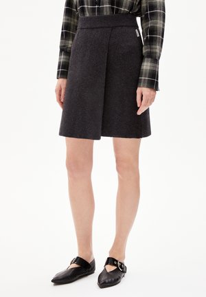 Femme portant une jupe portefeuille en laine gris foncé, une chemise noire à carreaux à manches longues et des ballerines noires pointues avec une bride à boucle.