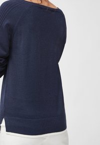 Personne portant un pull en maille bleu marine avec des détails côtelés sur les épaules et un pantalon blanc, vue de dos sur un fond uni.