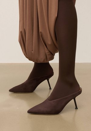 Mules de tacón alto de ante marrón con punta afilada y diseño elegante, combinados con medias marrón oscuro y tela marrón drapeada.