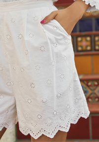Pantalones cortos blancos bordados con pequeños patrones florales, dobladillos en forma de onda y bolsillos laterales, elaborados con una tela ligera.