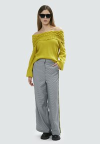 Maglione giallo a spalla scoperta con lavorazione a coste, abbinato a pantaloni larghi a quadri grigi con strisce gialle sui lati. Calzature nere e occhiali da sole.