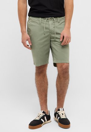 Man wearing olive green drawstring shorts, black and white sneakers, and a black shirt, standing against a plain gray background.