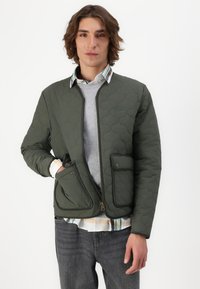 Grøn quiltet jakke med en frontlynlås, to forlommer og en høj krave, lagd over en grå sweater og en ternet skjorte.