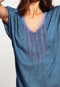 Niebieska tunika z krótkimi rękawami, z dekoltem w serek i ozdobnym, fioletowym oraz srebrnym haftem wzdłuż dekoltu, wykonana z teksturowanej tkaniny.