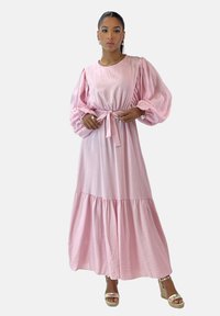 Vestido largo rosa con mangas abullonadas, cintura elástica y detalle de lazo. Bajo acampanado con textura fruncida. Combinado con sandalias de cuña doradas.